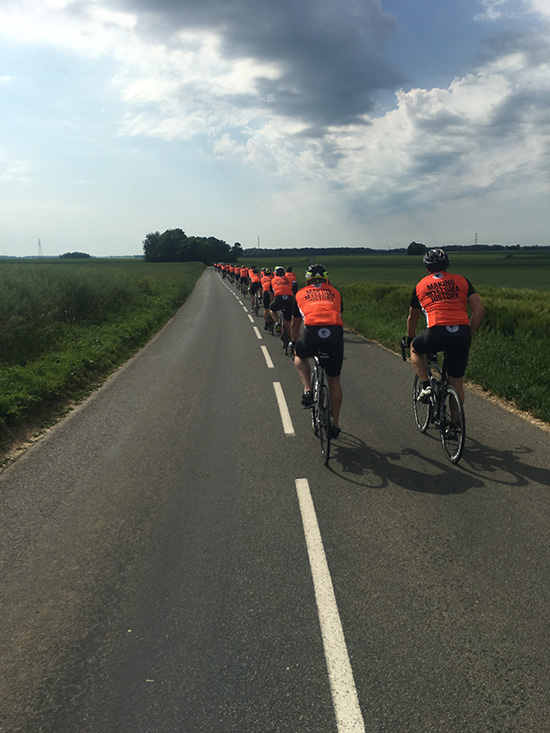 Single line of cyclists wearing orange myeloma uk t-shirts cycling along a narrow country lane